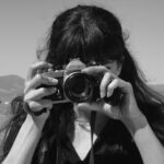 a black-and-white portrait of an anonymous woman taking a photo with an SLR camera with the desert in the background