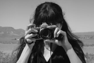 a black-and-white portrait of an anonymous woman taking a photo with an SLR camera with the desert in the background