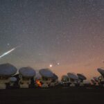 A bright light streaks across a starry, rosy-purple night sky on the left side of this image. An array of many radio telescopes are seen in the foreground.