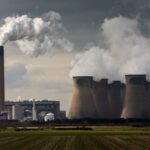 Sitting in a green field a power plant with smokestacks belches white smoke into the sky