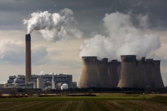 Sitting in a green field a power plant with smokestacks belches white smoke into the sky