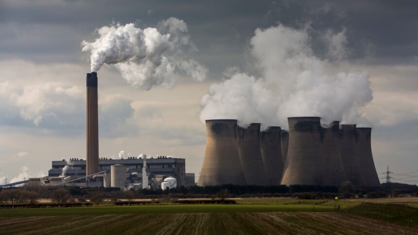 Sitting in a green field a power plant with smokestacks belches white smoke into the sky