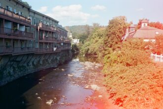 A film photo of a river near buildings with a sun spot on the right