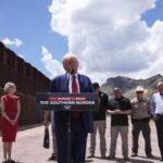 Donald Trump speaking behind a podium along the border wall in Arizona; the sky behind him takes up half the frame, and one woman dressed in red stands to his right, while a row men stand off to his left.