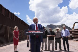 Donald Trump speaking behind a podium along the border wall in Arizona; the sky behind him takes up half the frame, and one woman dressed in red stands to his right, while a row men stand off to his left.