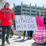 A man in a red sweater and glasses carries a Stop Lithium Mining sign which is also held by a woman in a pink shirt and skirt