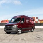 A maroon van sits in an empty lot in front of shipping containers