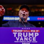 Elon Musk behind a podium with a Trump campaign sign speaks at a rally.