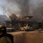 Firefighter in the foreground walks by a burning house in the background.