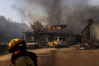 Firefighter in the foreground walks by a burning house in the background.