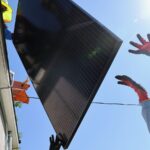 Two workers hand a solar panel to another worker on the roof a house.