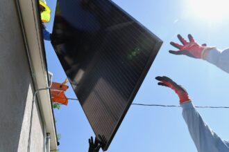 Two workers hand a solar panel to another worker on the roof a house.