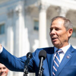 Photo of a man in a blue suit gesturing in front of a microphone in front of the Capitol