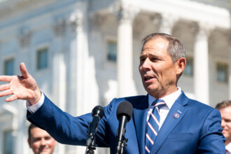 Photo of a man in a blue suit gesturing in front of a microphone in front of the Capitol