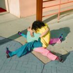 two people, one in a blue shirt and pink pants and the other in a yellow shirt and blue pants, embrace while seated on the sidewalk