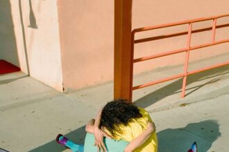 two people, one in a blue shirt and pink pants and the other in a yellow shirt and blue pants, embrace while seated on the sidewalk