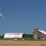 A windmill next to a barn with hay bales in front.