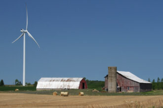 A windmill next to a barn with hay bales in front.