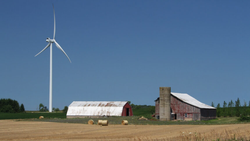 A windmill next to a barn with hay bales in front.