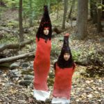 in the forest, two figures wear matching red garments reminiscent of dog stinkhorn mushrooms.