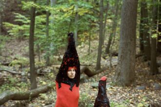 in the forest, two figures wear matching red garments reminiscent of dog stinkhorn mushrooms.