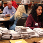 A woman sitting at a tabl;e covered in ballots. Other tables with ballots and people are visible in the background.