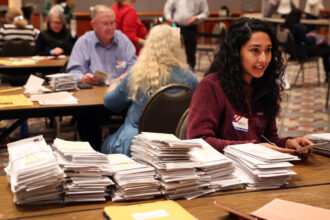 A woman sitting at a tabl;e covered in ballots. Other tables with ballots and people are visible in the background.