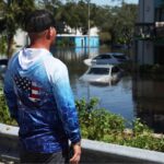 A person wearing an American flag hoodie surveys a flooded apartment complex and submerged parking lot in Clearwater, FL, after Hurricane Milton in 2024.