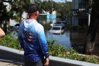 A person wearing an American flag hoodie surveys a flooded apartment complex and submerged parking lot in Clearwater, FL, after Hurricane Milton in 2024.