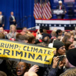 an african american person holds a sign up in the middle of a crowd of people. The sign is a fabric banner that says TRUMP: CLIMATE CRIMINAL.. Behind her is a blurry background showing a man in a hat at a podium and an audience with their phones out looking away from the podium.