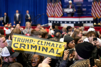 an african american person holds a sign up in the middle of a crowd of people. The sign is a fabric banner that says TRUMP: CLIMATE CRIMINAL.. Behind her is a blurry background showing a man in a hat at a podium and an audience with their phones out looking away from the podium.