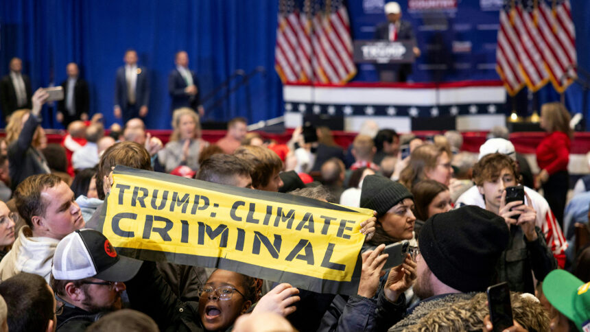 an african american person holds a sign up in the middle of a crowd of people. The sign is a fabric banner that says TRUMP: CLIMATE CRIMINAL.. Behind her is a blurry background showing a man in a hat at a podium and an audience with their phones out looking away from the podium.