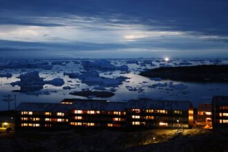 Icebergs float in Disko Bay, near a row of warmly illuminated houses, as moonlight shines through the clouds on September 03, 2021 in Ilulissat, Greenland.