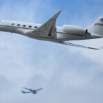 A small private jet flies through a blue sky with a passenger jet in the background