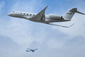 A small private jet flies through a blue sky with a passenger jet in the background