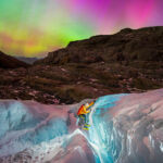 a glacier hiker climbs in the foreground with a headlamp as a rainbow colored aurora dances above