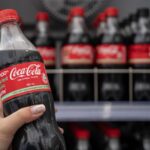 Close-up of a hand holding a Coca-Cola bottle, with a shelf of more bottles in the background.