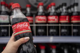 Close-up of a hand holding a Coca-Cola bottle, with a shelf of more bottles in the background.