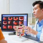 A male doctor sits next to a computer screen with diagnostic images while gesturing at a 3D model of a colon, across the desk from an older man wearing an orange shirt