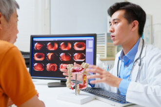 A male doctor sits next to a computer screen with diagnostic images while gesturing at a 3D model of a colon, across the desk from an older man wearing an orange shirt