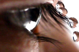 A closeup shows an eye framed by eyelashes, with four droplets of water pictured landing on an eyelash, sliding off, and falling.