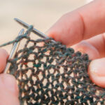 A close-up view of fingertips holding silver knitting needles, with light shining through the loose stitches in the cast fabric