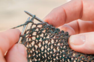 A close-up view of fingertips holding silver knitting needles, with light shining through the loose stitches in the cast fabric