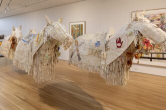 an installation of textile horses is suspended in a gallery with works on the wall and pedestal in the background