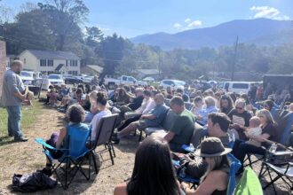 Outside a brick building, under a blue sky with mountains in the background, a man in a grey shirt and jeans holding a hat stands in front of a group of people seated in folding chairs.