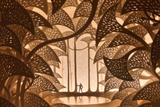 an intricate papercut world with a tiny figure wandering through tall trees. the piece is backlit by warm yellow light