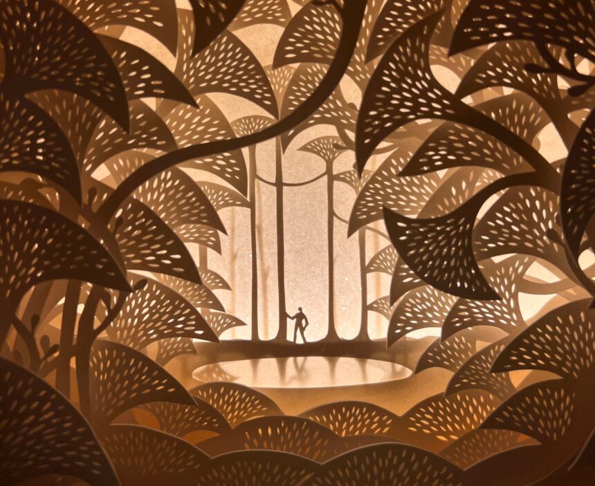 an intricate papercut world with a tiny figure wandering through tall trees. the piece is backlit by warm yellow light