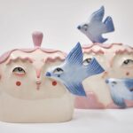 two ceramic sculptures of cute heads and faces with pink hair, with bluebirds flying around them