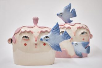 two ceramic sculptures of cute heads and faces with pink hair, with bluebirds flying around them