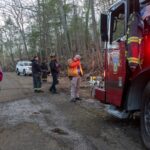 Firefighters mill around on a dirt road next to a red firetruck that says Volunteer on the side